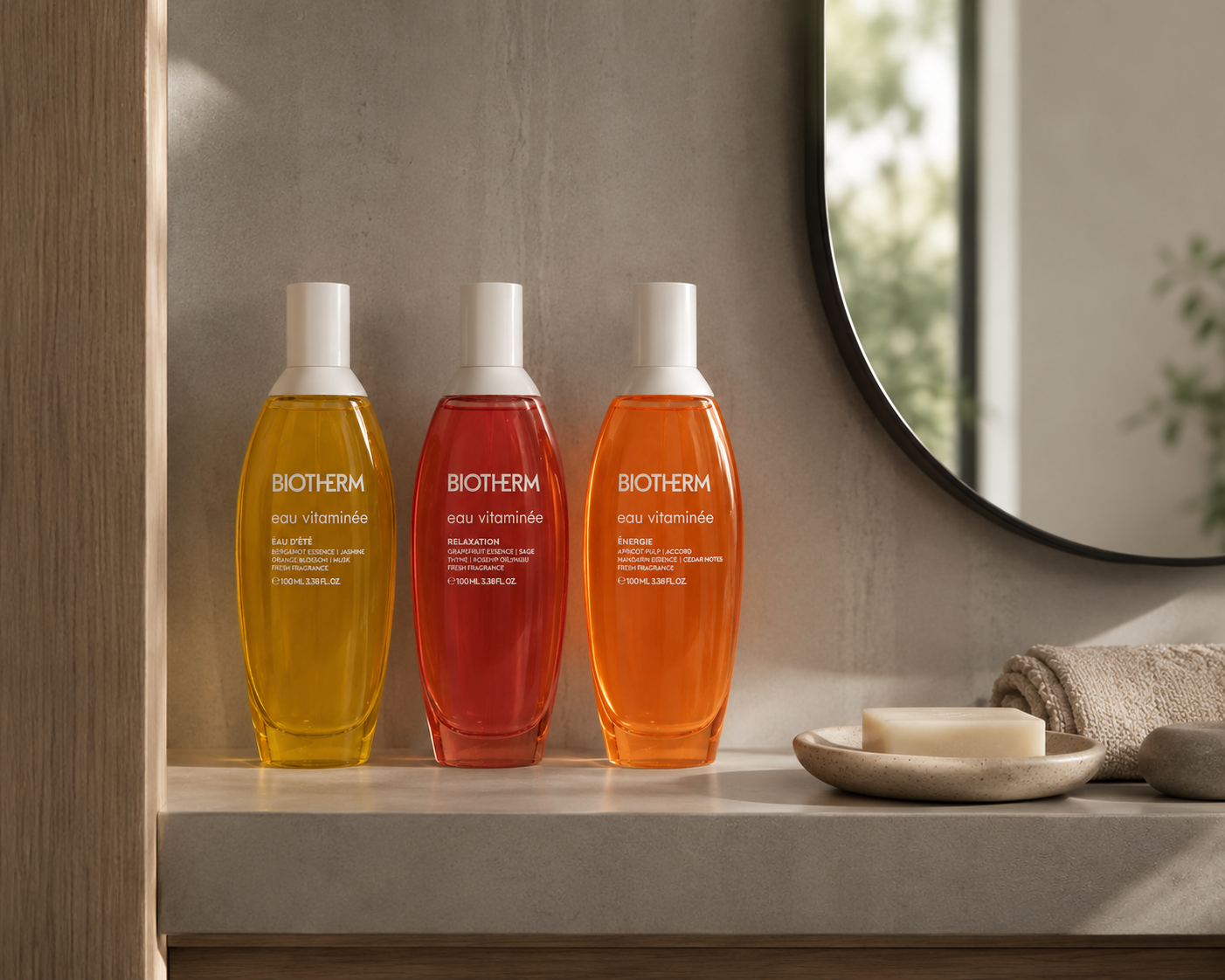 Drei farbige Biotherm Eau Vitaminée Flakons stehen auf einer modernen Badezimmerablage neben einem runden Spiegel, Seifenschale und Handtuch in warmem Tageslicht.