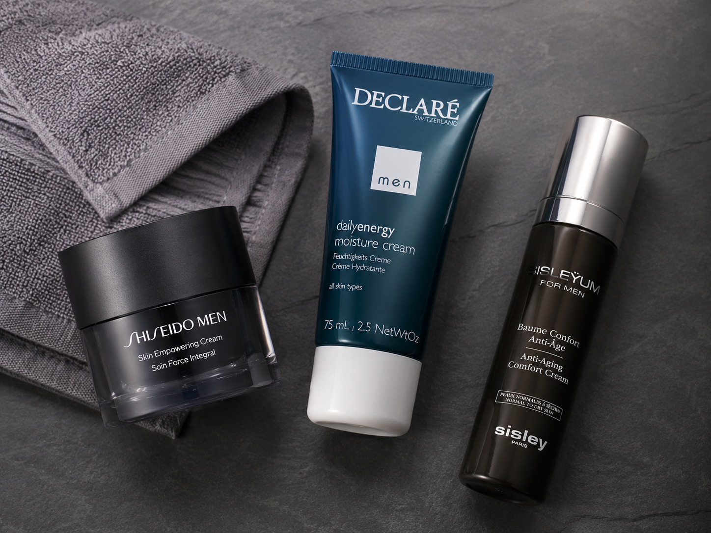 Herren-Gesichtspflege auf Schiefer: Shiseido Men Skin Empowering Cream, Declaré Men Dailyenergy   Feuchtigkeitscreme und Sisley Anti-Aging Comfort Cream neben einem grauen Handtuch