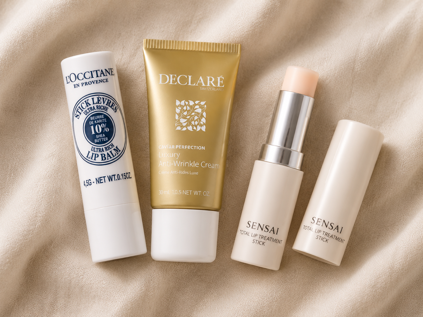 Lippenbalsam von L'Occitane, Antifaltencreme von Declaré und ein Lippenpflegestift von Sensai als Flatlay auf beigefarbenem Stoff.