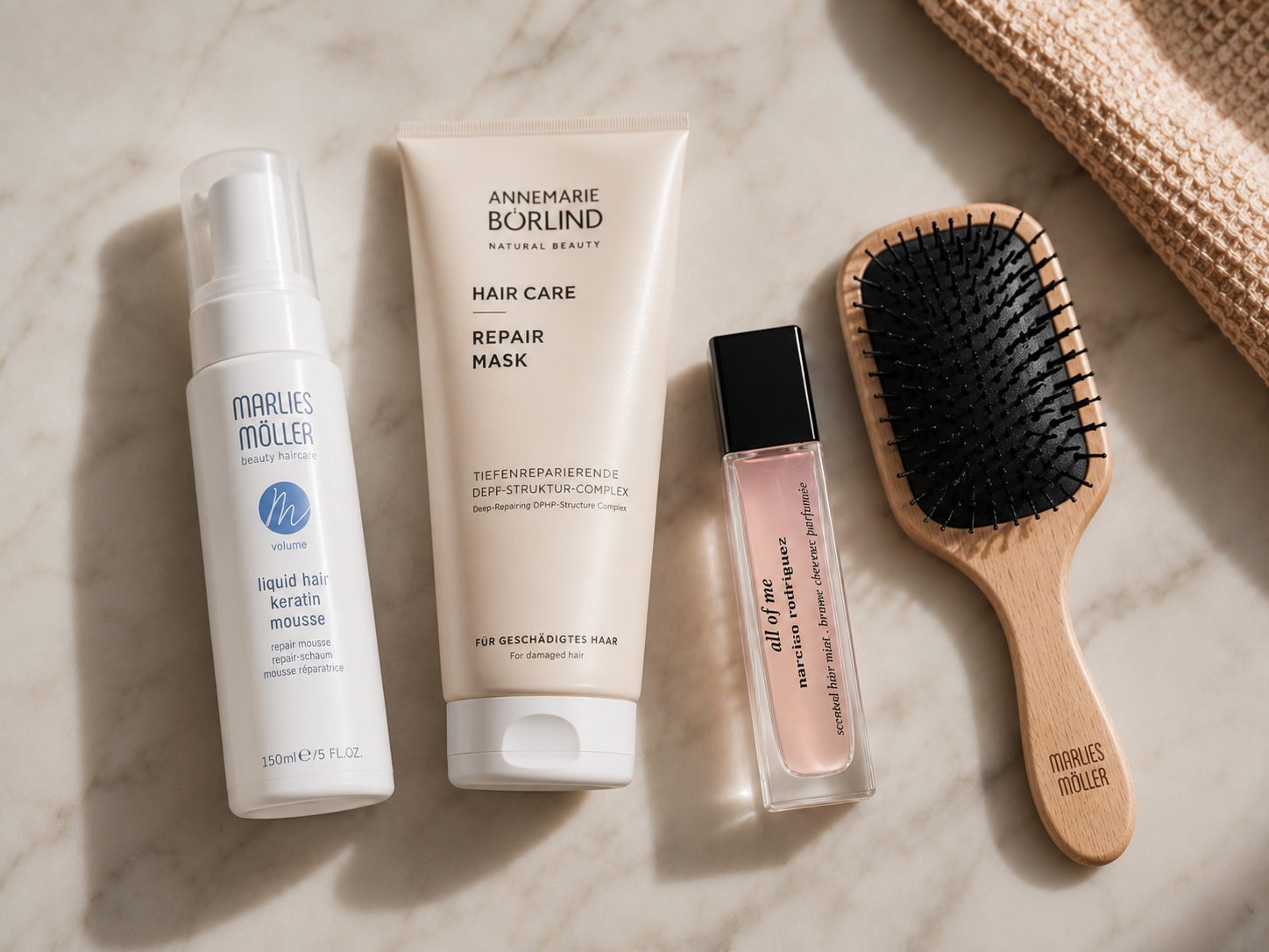 Haarpflegeprodukte von Marlies Möller und Annemarie Börlind, ein Narciso Rodriguez Haarparfüm und eine Holzhaarbürste als Flatlay auf Marmor.