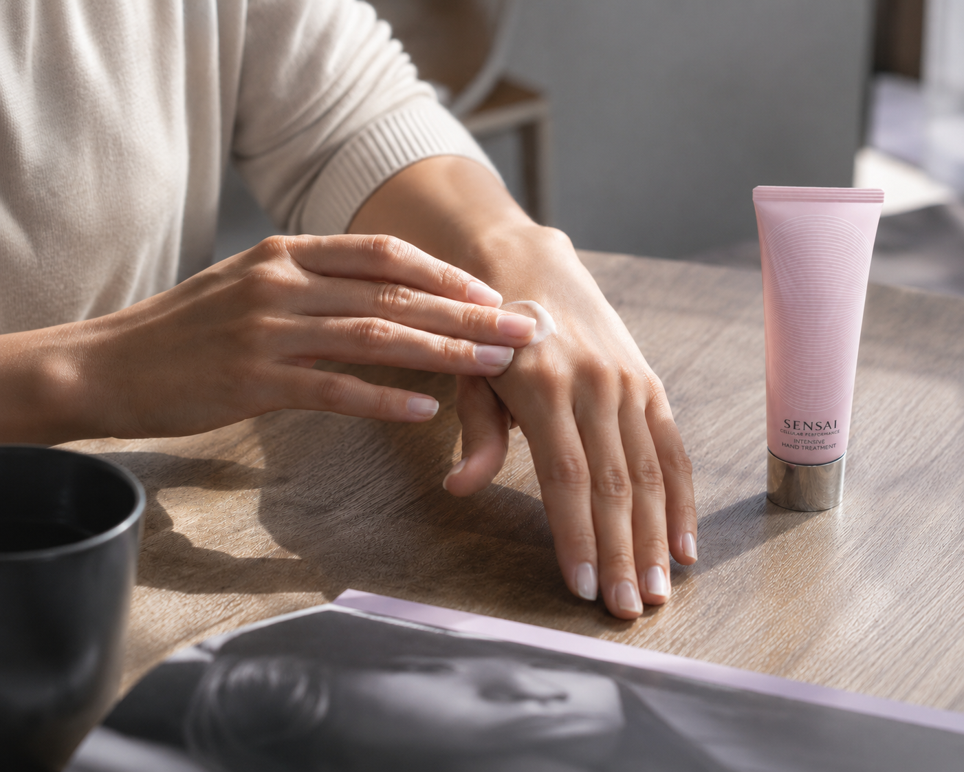Eine Person cremt sich am Holztisch die Hände ein; daneben steht eine rosa Tube Sensai „Intensive Hand Treatment.