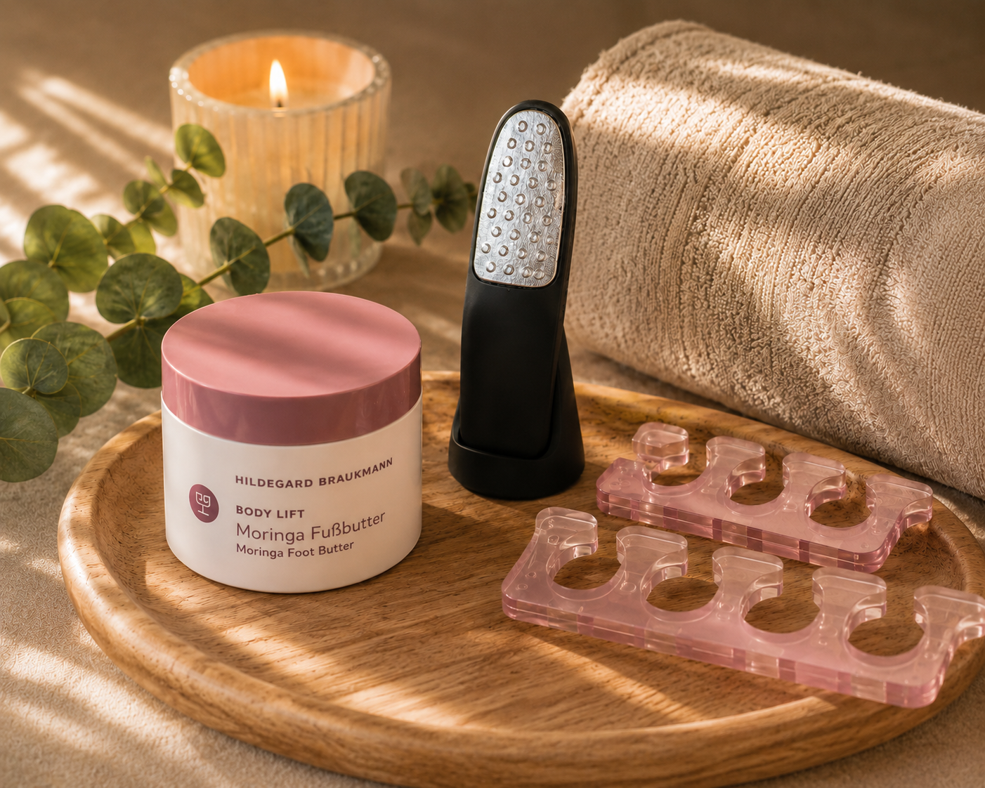 Ein Tiegel Hildegard Braukmann „Moringa Fußbutter“, eine elektrische Hornhautfeile und rosa Zehentrenner auf einem Holztablett neben Handtuch und brennender Kerze.