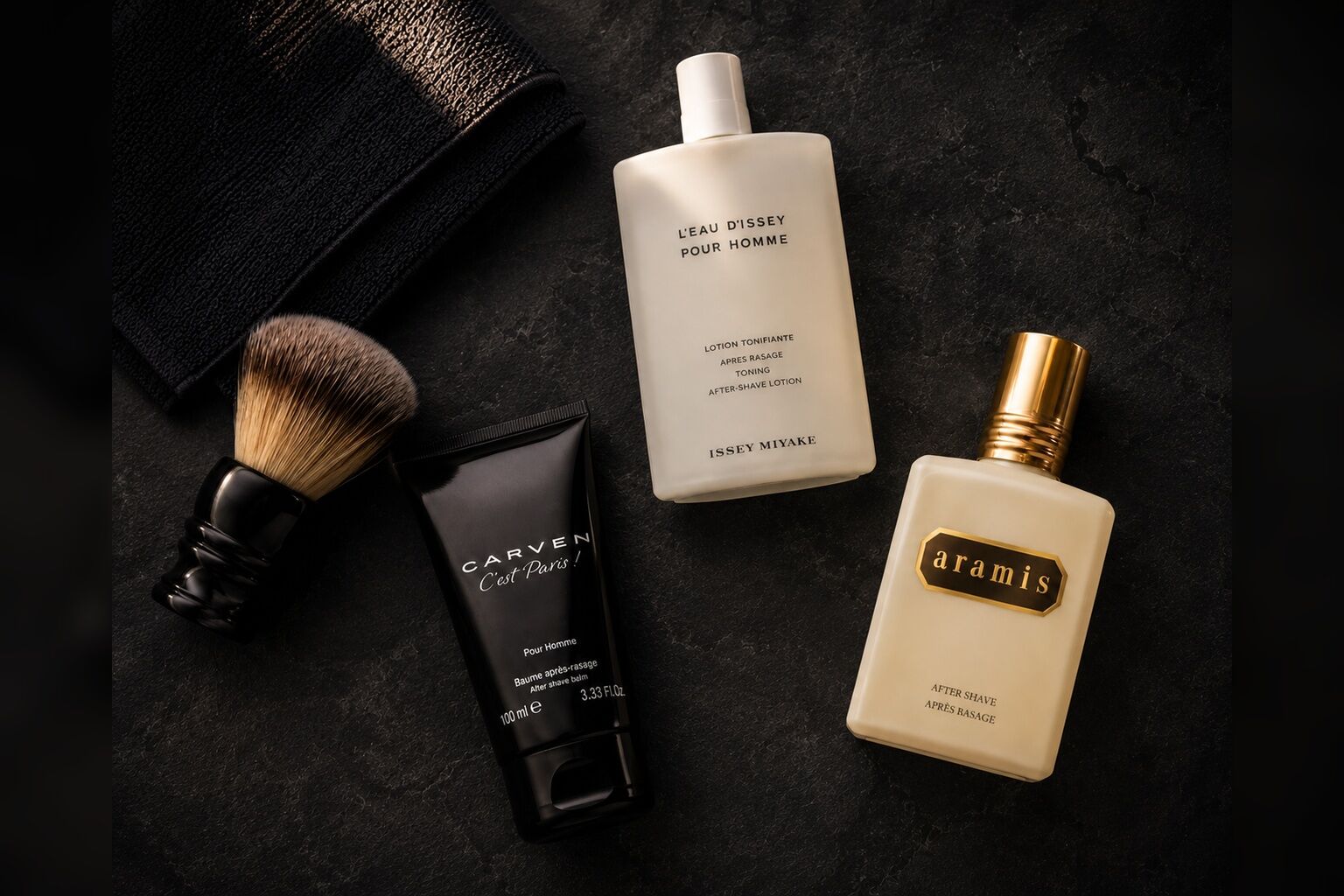 Aftershave-Produkte von Carven, Issey Miyake und Aramis auf dunklem Stein, arrangiert mit Rasierpinsel und schwarzem Handtuch in warmer, maskuliner Lichtstimmung.