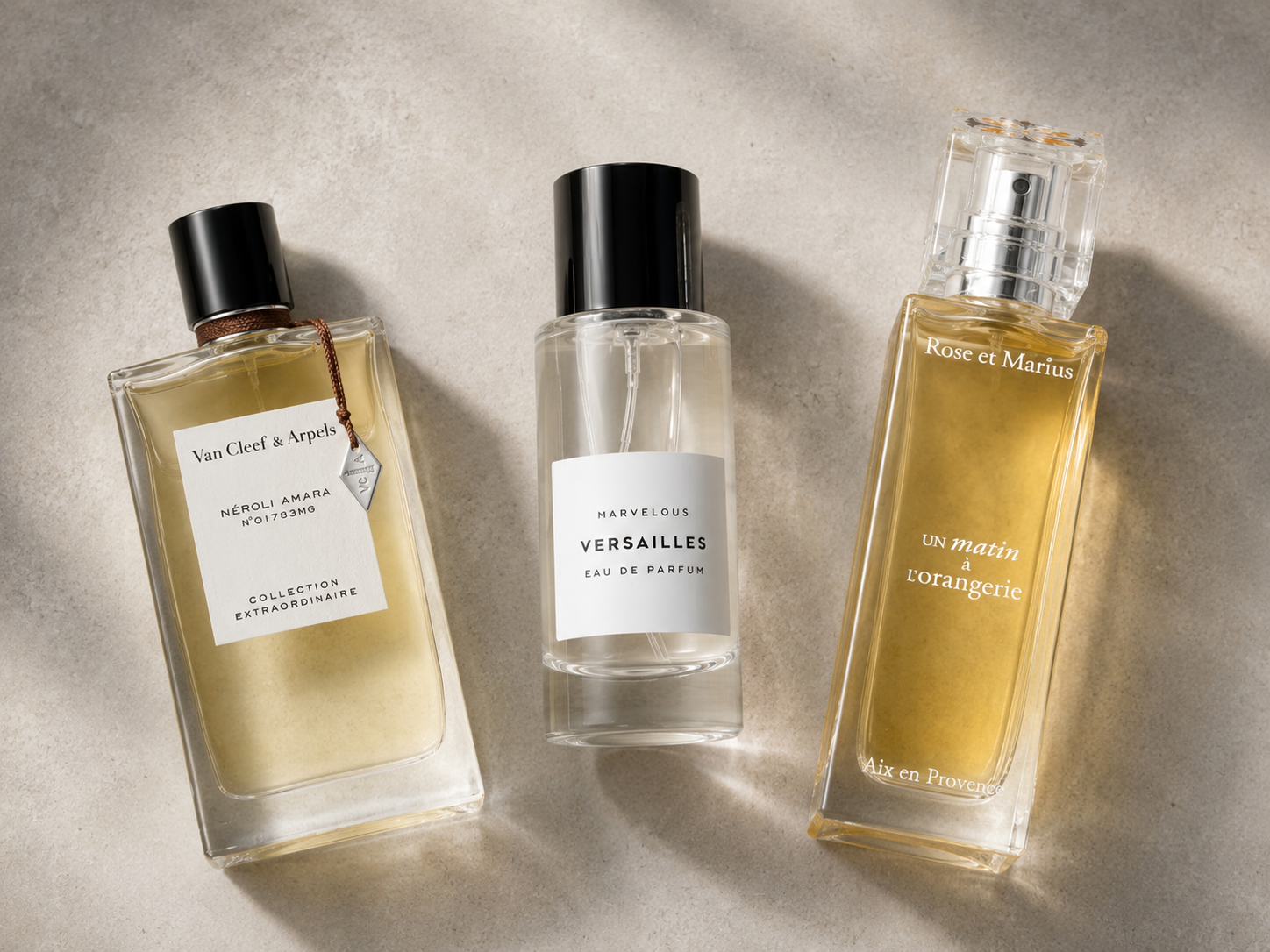 Flatlay mit drei Unisex-Parfums auf hellem Sandstein: Van Cleef & Arpels Néroli Amara, Marvelous   Versailles und Rose et Marius Un matin à l'orangerie