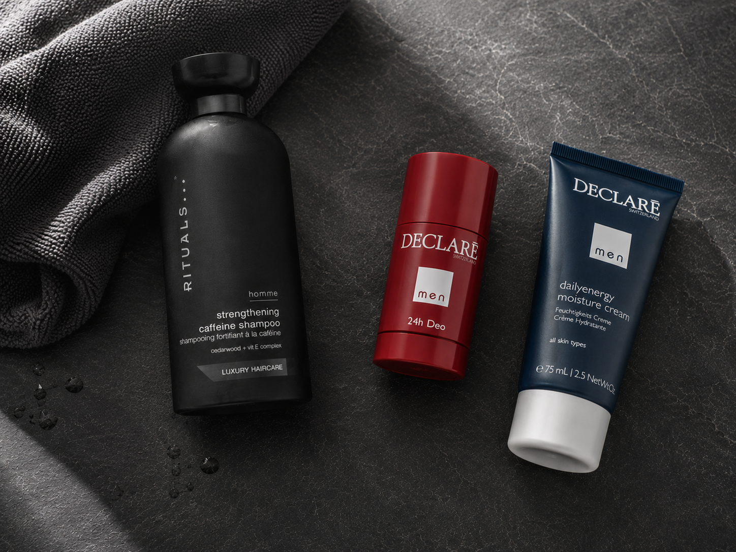 Herren-Körperpflege in der Aufsicht: Rituals Homme Caffeine Shampoo, Declaré Men 24h Deo und   Declaré Men Dailyenergy Feuchtigkeitscreme auf dunklem Schiefer