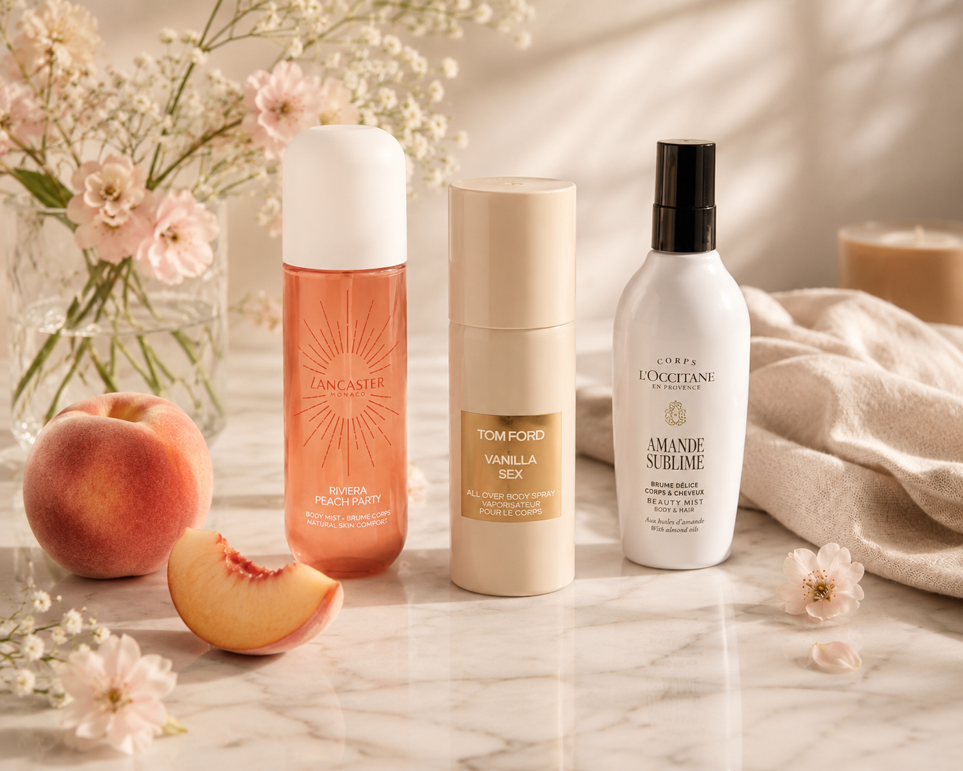 Drei luxuriöse Body Sprays von Lancaster, Tom Ford und L’Occitane auf heller Marmorfläche, dekoriert mit Pfirsich, zarten Blüten und weichem Leinen in warmer, natürlicher Lichtstimmung.