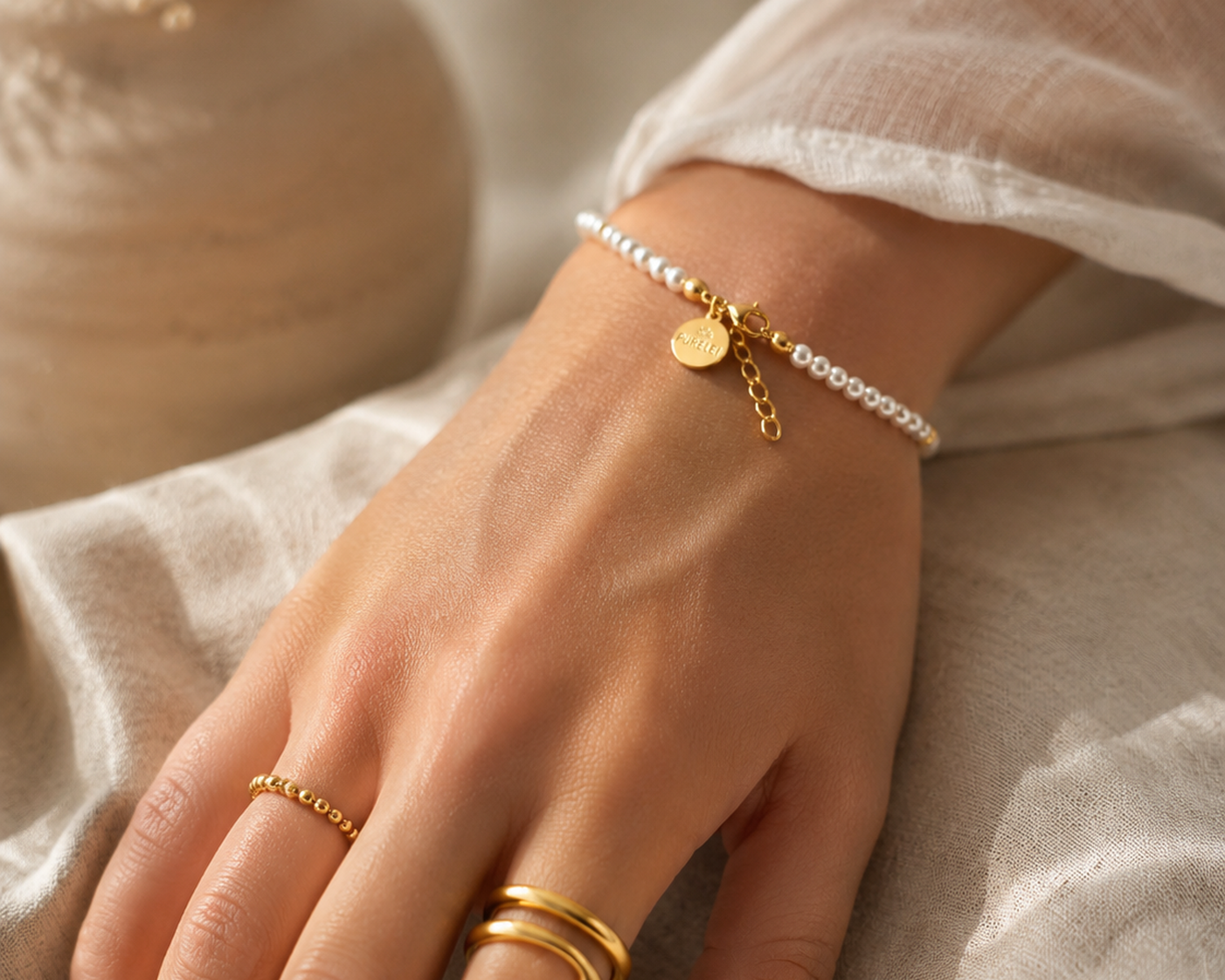 Hand mit Purelei Perlenarmband, goldenem Perlenring und gestapelten schlichten Goldringen auf   hellem Leinenstoff