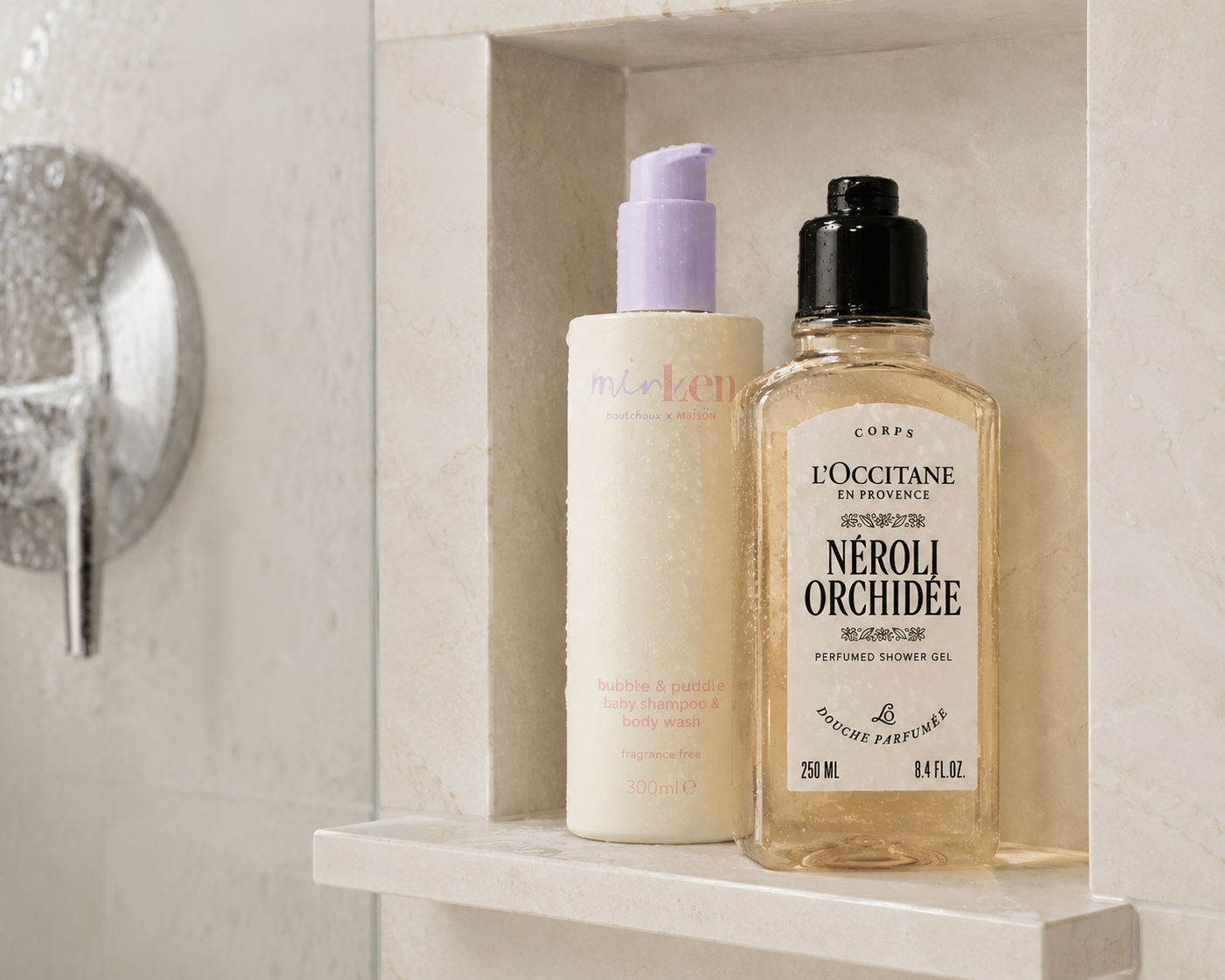 Ein Baby-Shampoo von Minois und ein L'Occitane „Néroli & Orchidée“ Duschgel nebeneinander in einer hellen Naturstein-Duschablage.